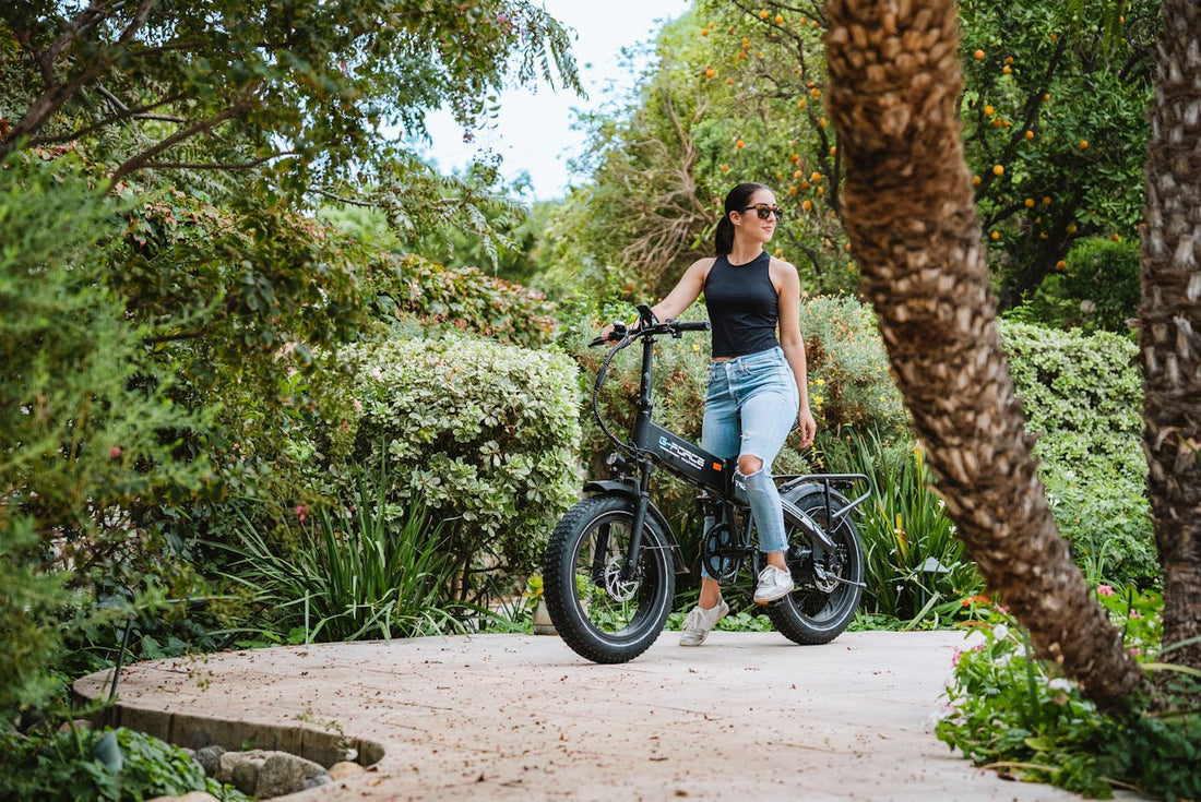 Femeie cu ochelari de soare lângă o bicicletă electrică pliabilă, cu roți groase, pe o alee dintr-o grădină verde